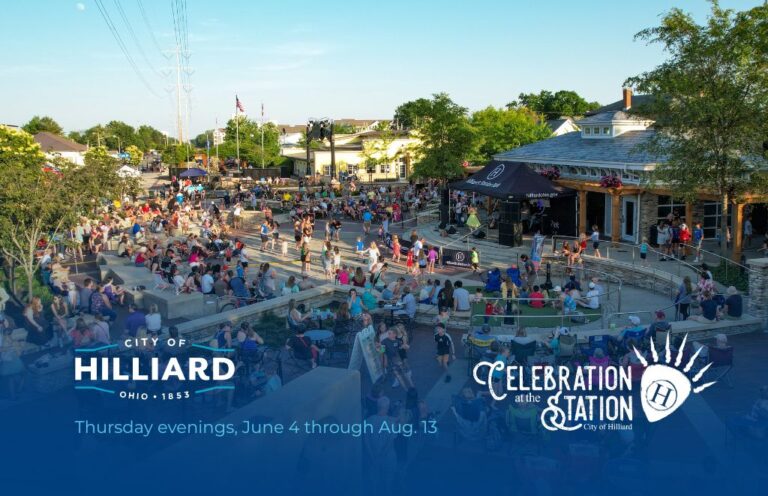 Outdoor evening festival in a town square with a stage and many seated attendees, banners say Hilliard celebration at the station.