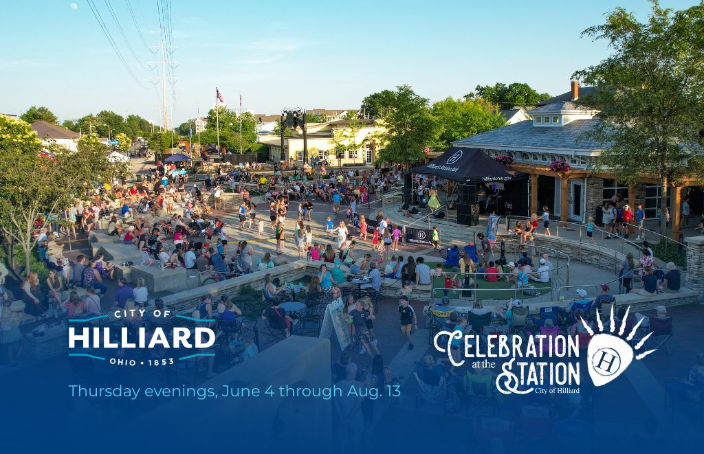 Outdoor evening festival in a town square with a stage and many seated attendees, banners say Hilliard celebration at the station.